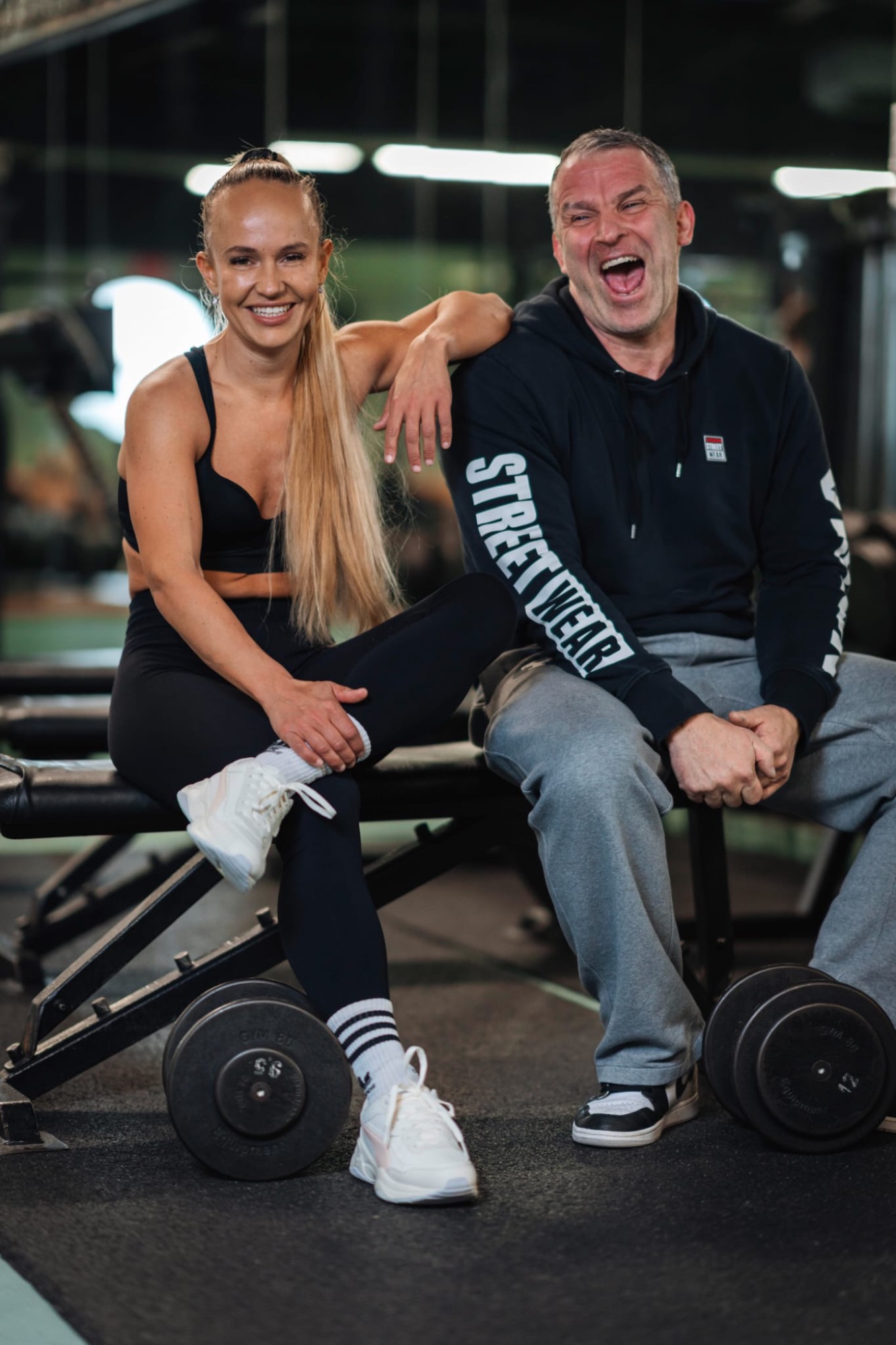 Lächelnde Sportlerin und Trainer sitzen neben Hanteln im Fitnessstudio.